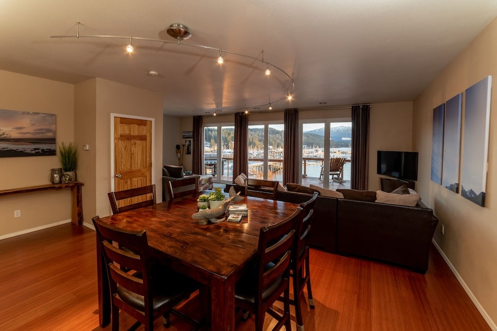 The open-concept living area features a wooden dining table surrounded by dark chairs. Large windows provide views of Auke Bay Harbor, allowing ample natural light to fill the room. A cozy seating area with a television is visible in the background.