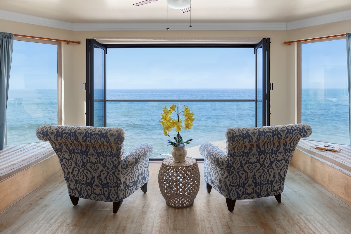 Two comfortable armchairs are positioned facing large open windows, offering a direct view of the Pacific Ocean. A small, decorative table sits between the chairs, adorned with vibrant yellow flowers. Natural light fills the space, enhancing the serene atmosphere.