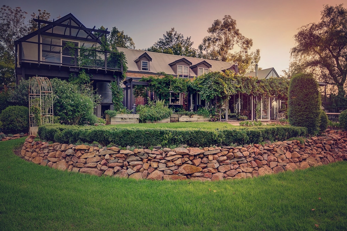 A charming historic house is framed by lush greenery and flowering vines, with a stone-lined garden featuring manicured hedges. The front porch offers a welcoming spot for relaxation, and large windows reflect the serene surroundings, enhancing the property's inviting exterior.
