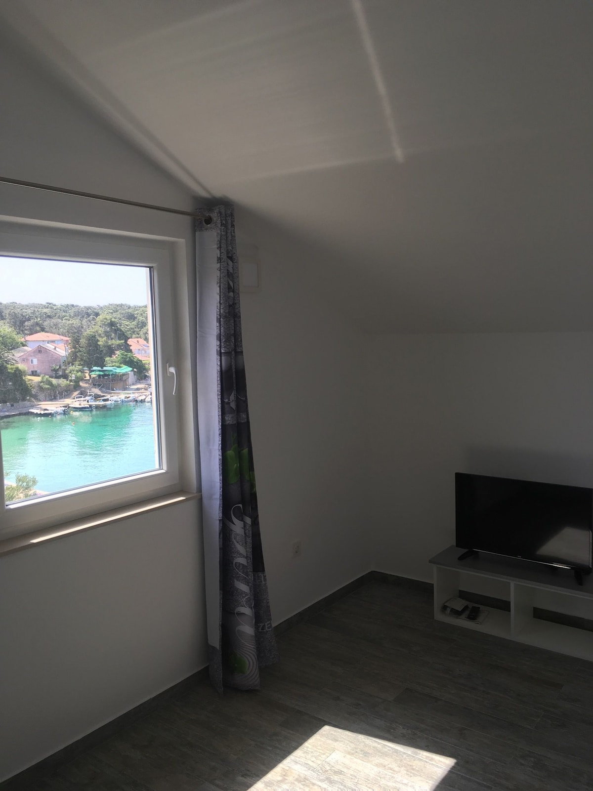 A well-lit corner of a room features a large window offering a view of the tranquil sea and surrounding landscape. A flat-screen television sits on a simple white stand, complemented by light-colored walls and flooring, creating a bright and airy atmosphere.