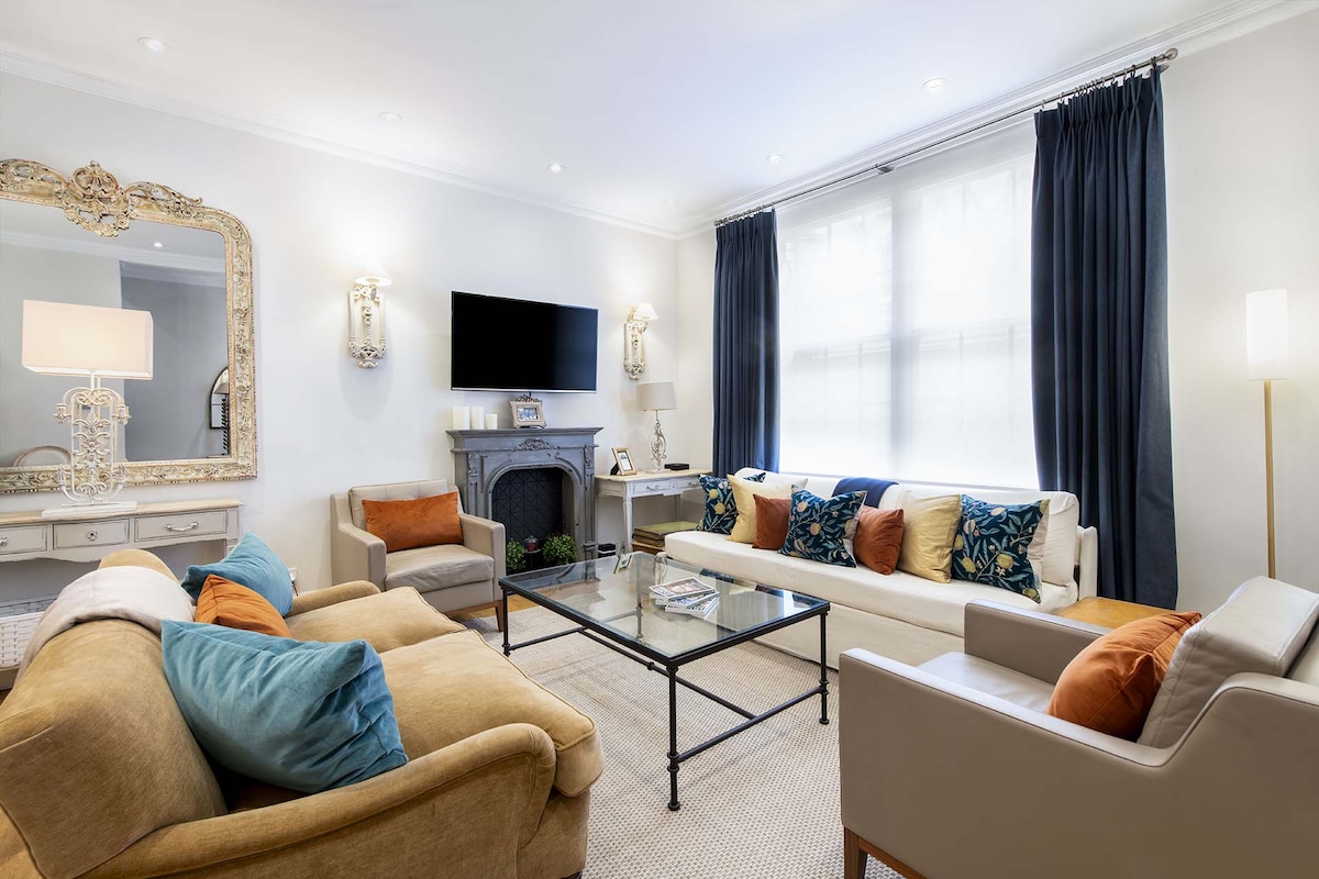 The open plan living area features a mix of seating options including a large comfortable sofa and two armchairs. A glass coffee table occupies the center, with decorative cushions adding colors to the space. A modern television is mounted on the wall, and natural light filters through the elegant curtains.