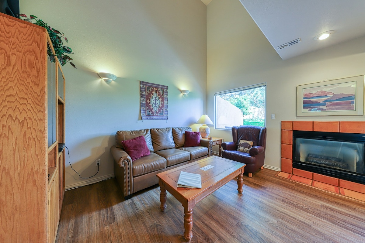A cozy living area is presented with a comfortable brown leather sofa and an armchair, featuring decorative cushions. A wooden coffee table is centered between the seating, and natural light enters through a window adorned with minimal coverings. A fireplace and wall art add warmth to the space.