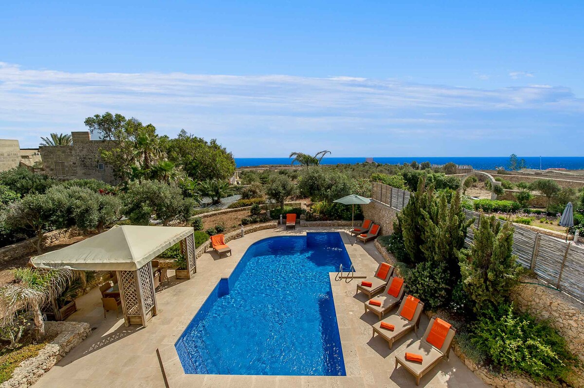A spacious outdoor area features a large swimming pool surrounded by eight sun loungers with orange cushions. Two shaded areas provide relief from the sun, and lush greenery complements the scenic views of the sea and garden.