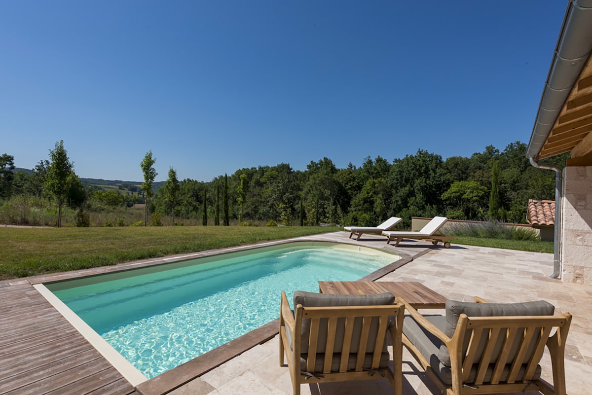 An outdoor swimming pool is surrounded by sun loungers, set against a backdrop of lush greenery. The clear water reflects the bright blue sky, while a spacious wooden deck offers ample room for relaxation and leisure.