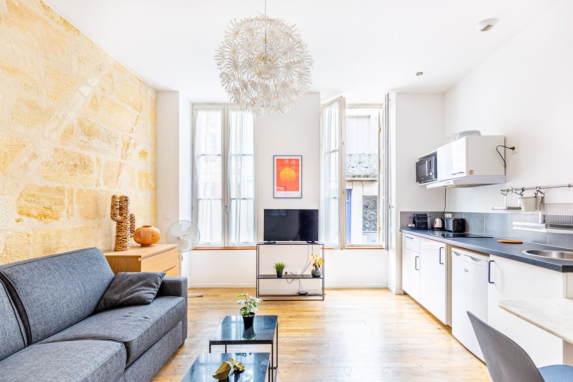 A bright living area features large windows with open shutters, flooding the space with natural light. A comfortable gray sofa is positioned next to a glass coffee table. A mounted television is visible across from the sofa, with a well-equipped kitchenette nearby, complete with modern appliances.