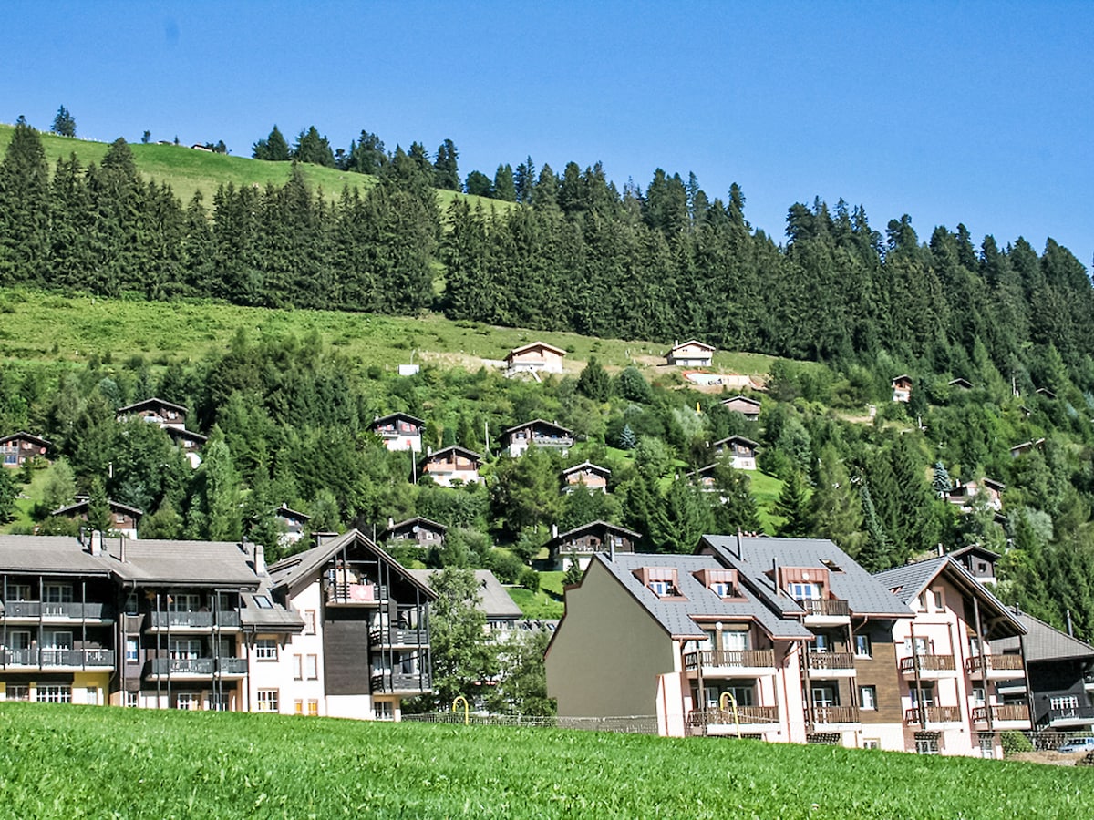 The image showcases a hillside view, featuring a blend of residential buildings nestled among lush greenery. The foreground includes a cluster of multi-story apartments, while the background displays trees and additional homes ascending the hill.