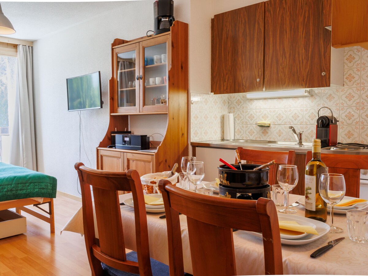 A well-appointed studio apartment features a dining table set for four, complete with glassware and cutlery. In the background, a compact kitchenette showcases modern appliances, while a comfortable sleeping area with green bedding is visible. A television is mounted on the wall, and natural light enters through a nearby window.