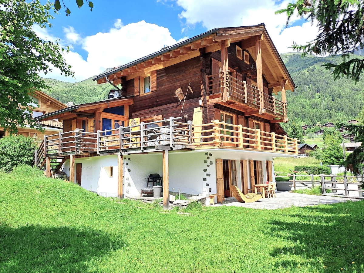 A two-story wooden house is set amid lush greenery, featuring multiple balconies and a spacious terrace. The ground floor displays stone and wooden elements, while the upper level is adorned with a rustic facade. Surrounding trees enhance the tranquil setting, with mountain views in the background.