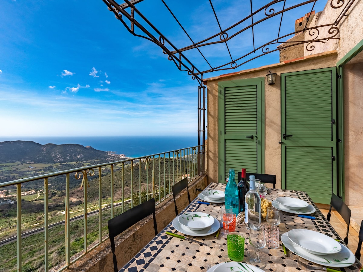 The terrace showcases a dining table set for several guests, adorned with plates and glasses. Lush greenery and a panoramic ocean view extend in the background, framed by green shutters, under a structured overhead canopy.
