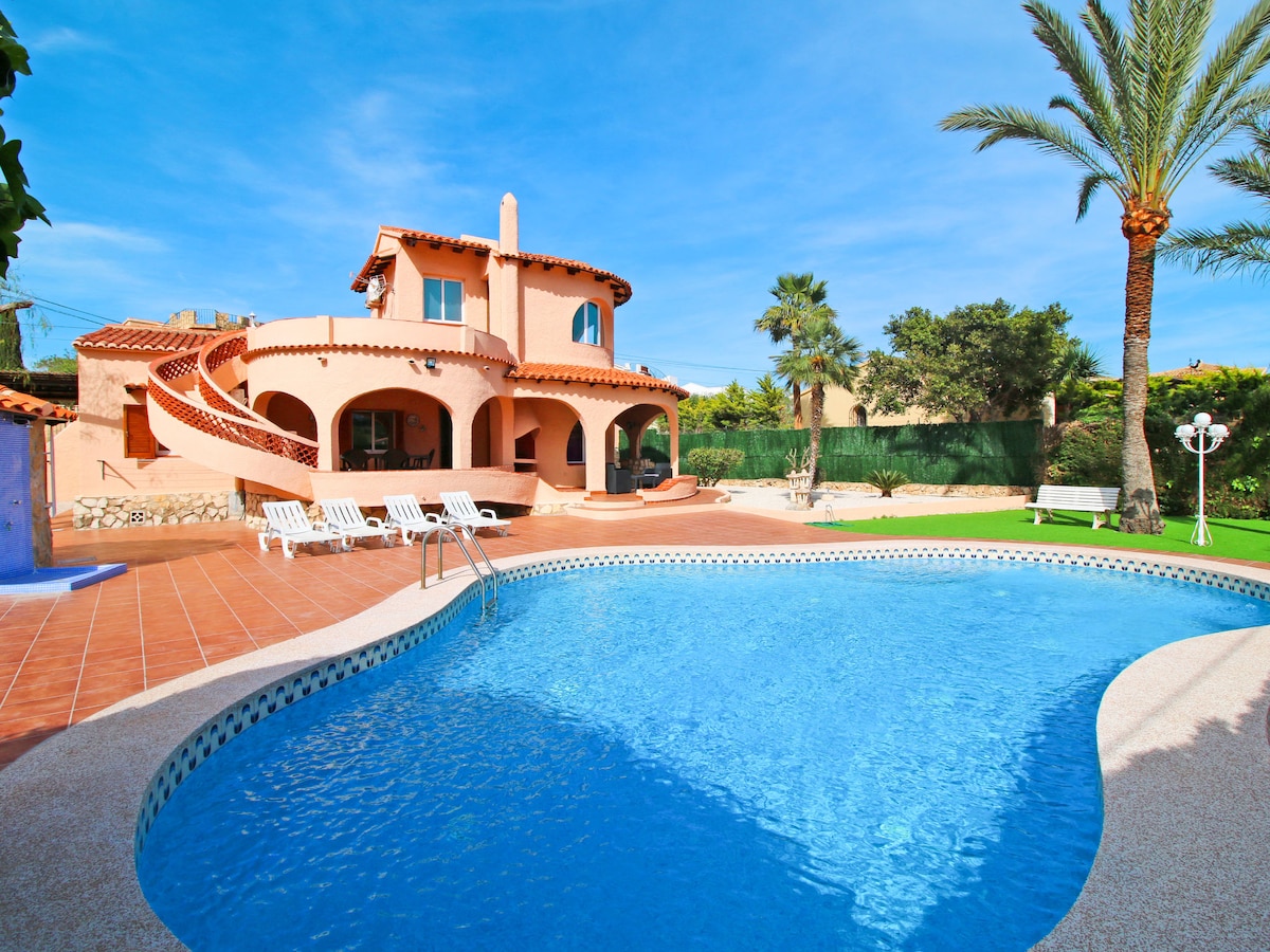 A kidney-shaped swimming pool is highlighted in the foreground, surrounded by a tiled deck with sun loungers. The villa features a pink exterior with a spiral staircase leading to the upper level. Lush palm trees and greenery create a private outdoor setting.