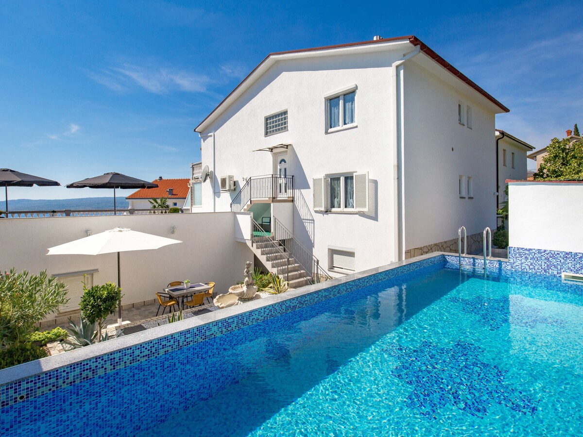 A white apartment building is seen beside a bright blue swimming pool. Sun loungers and umbrellas are positioned around the pool area, complemented by a seating arrangement featuring a table and chairs. The ocean is faintly visible in the background under a clear blue sky.