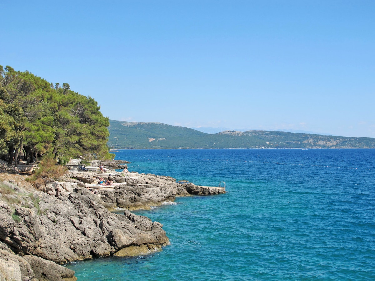 The image depicts a rocky coastline meeting the clear blue sea. Lush greenery is present along the shore, with a distant mountainous landscape visible under a bright sky. The calm water reflects shades of turquoise and deep blue, inviting leisurely outdoor activities.