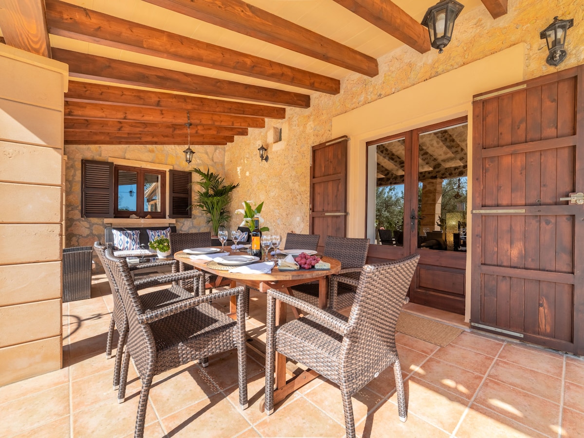 An outdoor dining area is displayed under a wooden-beamed ceiling, featuring a round table surrounded by wicker chairs. A set of glasses and a decorative fruit bowl are set on the table, with doors leading into the interior visible in the background.