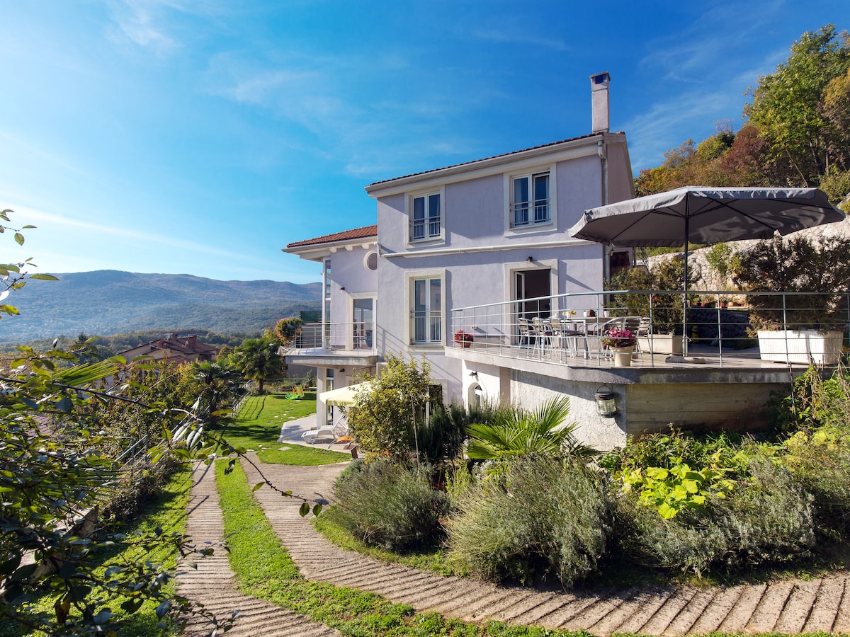 The exterior of the property features a two-story house surrounded by gardens and trees. A spacious terrace with outdoor furniture is visible, offering views of the surrounding hills and blue sky. Pathways meander through well-maintained landscaping, creating an inviting atmosphere.