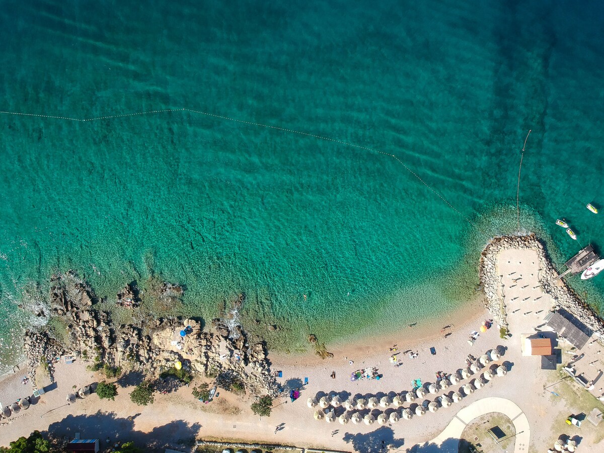 An aerial view of a beach reveals clear turquoise waters gently lapping against a sandy shore. Sun loungers and umbrellas line the beach, while a rocky area is visible nearby. The scene captures a peaceful seaside setting, ideal for relaxation.