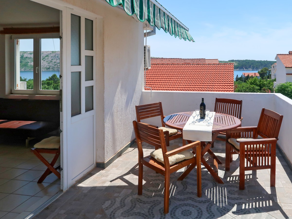A balcony is visible featuring a wooden table surrounded by four chairs, each with cushions. A bottle and two glasses are positioned on the table. The area is partially shaded with a striped awning, while a view of the sea and surrounding landscape can be seen in the distance.