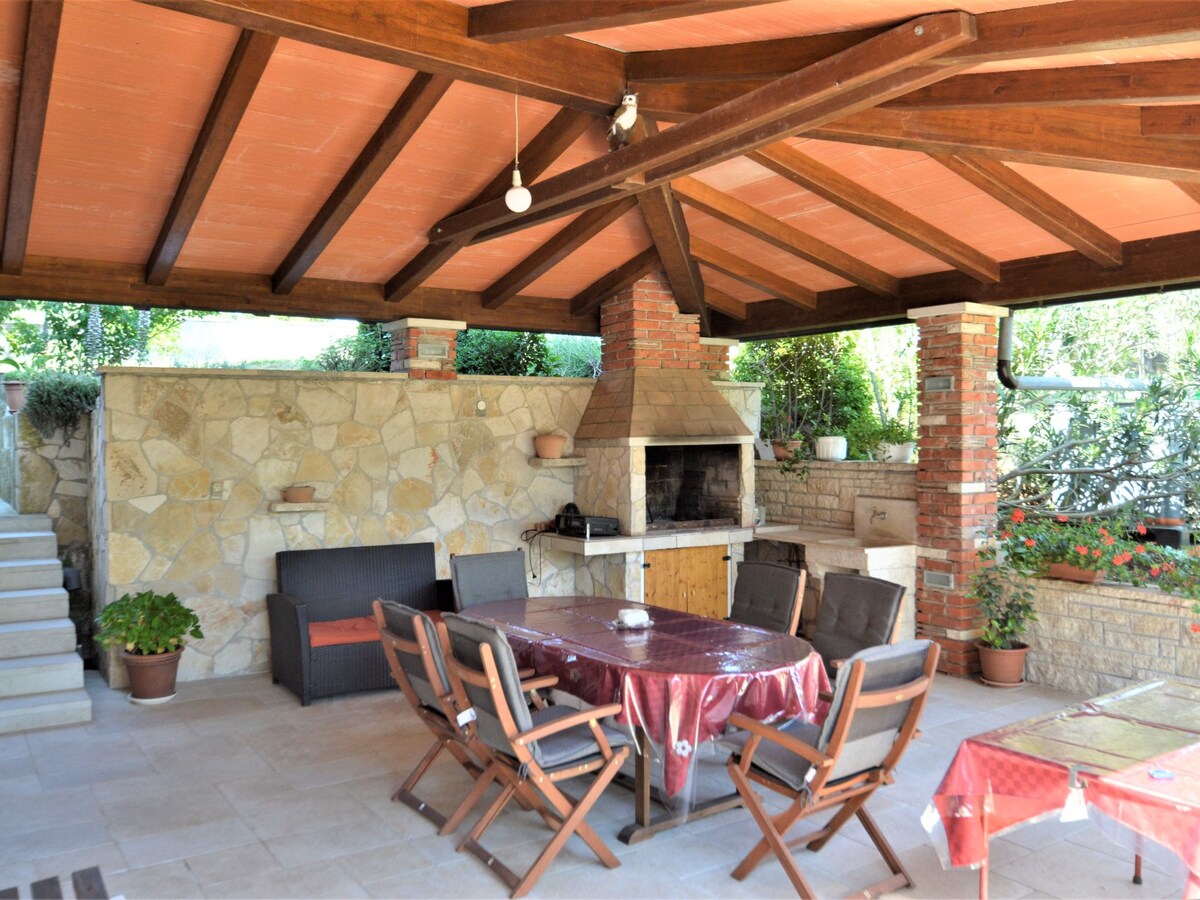 An outdoor terrace is sheltered by a wooden roof with exposed beams. A stone barbecue is featured alongside ample seating, including a large table surrounded by wooden chairs. Green plants add life to the stone walls, providing a serene atmosphere.