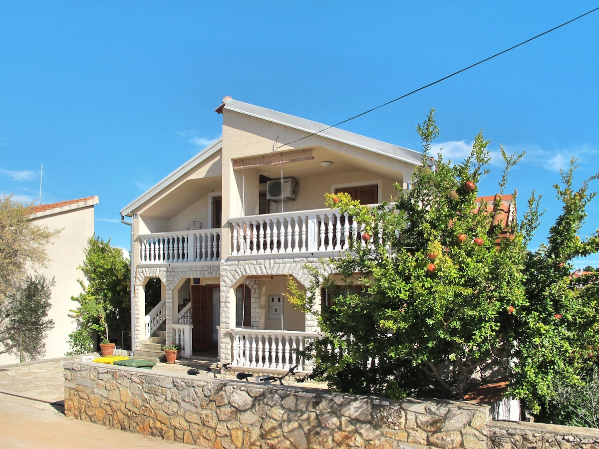 A two-story house is presented with a distinct architectural style, featuring a covered balcony with decorative railings. Lush greenery surrounds the entrance, and potted plants add a touch of color. The exterior walls are light-colored, complementing the clear blue sky above.