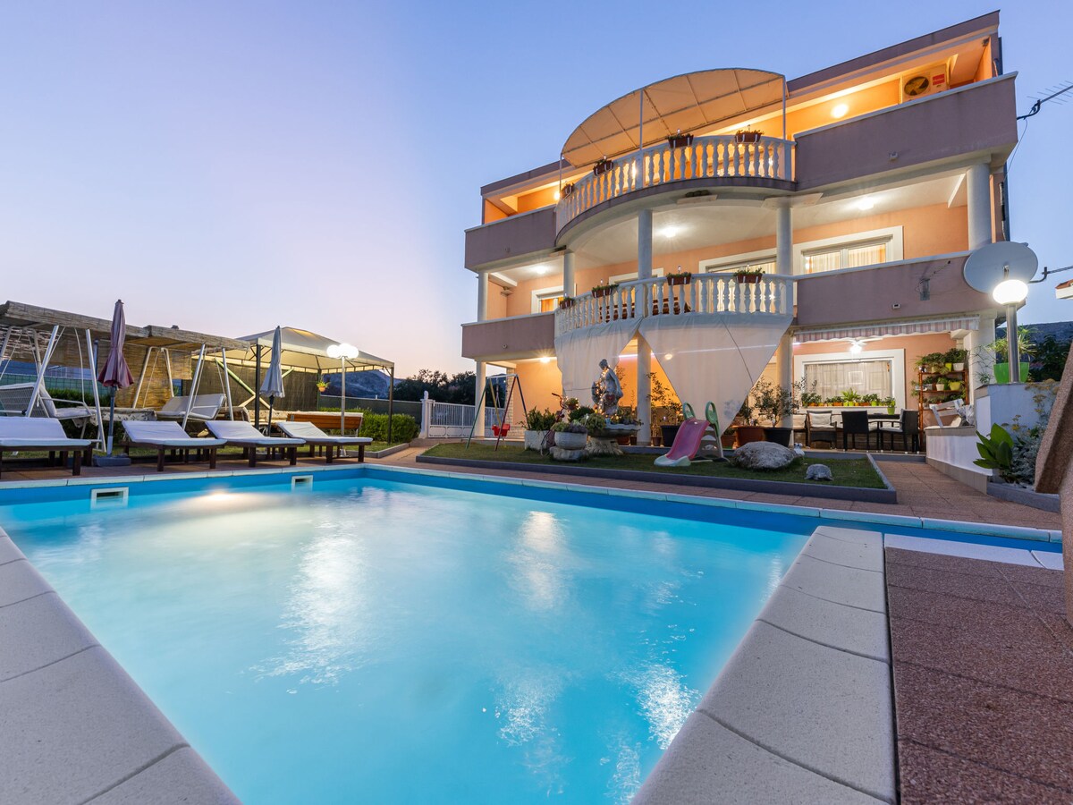 A multi-story villa features a spacious swimming pool surrounded by loungers and shaded areas. The exterior showcases a covered terrace, and balconies offer scenic views. Soft evening light enhances the atmosphere, illuminating the architectural details of the building.