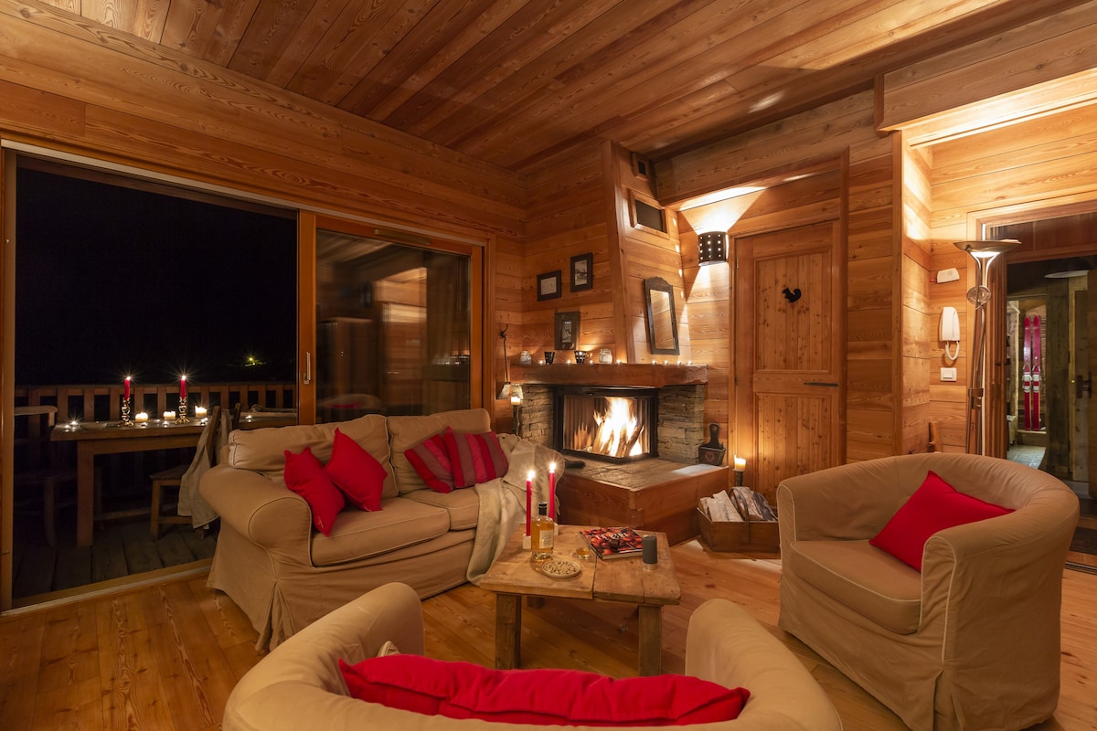 A warmly lit living area features wooden paneling and cozy seating arrangements with plush cushions. A stone fireplace serves as the focal point, offering warmth and ambiance. Large glass doors provide a view of the outdoor terrace, enhancing the inviting atmosphere.