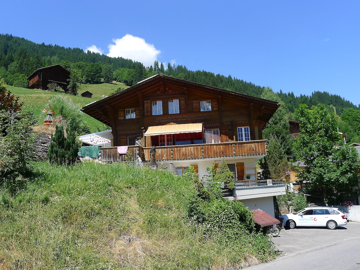 The chalet is nestled on a slope, showcasing a wooden façade and an inviting balcony. Surrounded by trees and meadows, the bright blue sky contrasts with the lush greenery, enhancing the tranquil setting. A parking area is visible near the entrance.