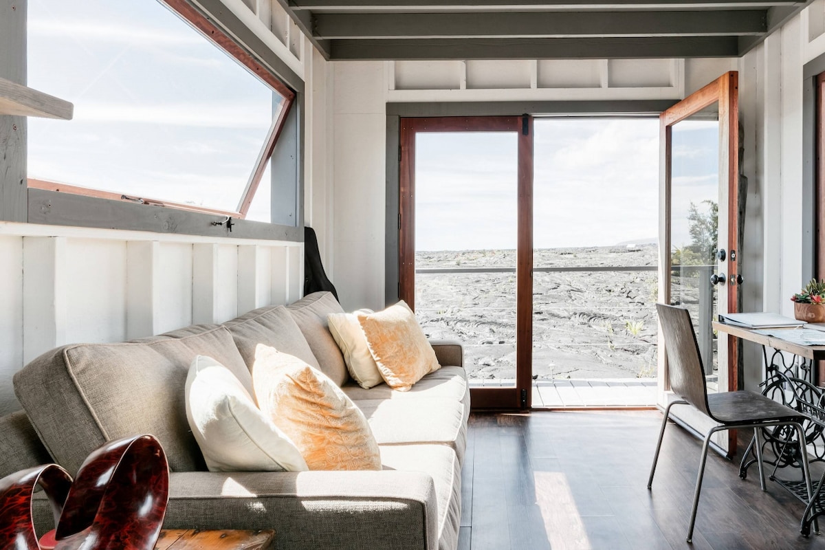 A cozy living area is highlighted by a comfortable couch adorned with cushions, facing large doors that open to reveal stunning views of the surrounding lava landscape. Natural light streams in, creating a bright atmosphere, complemented by a small dining table nearby.