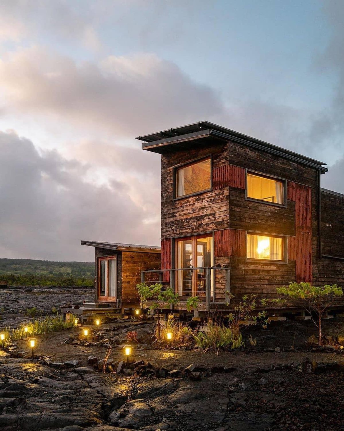 A modern tiny house features wooden exterior panels, illuminated by warm light from its large windows. Surrounding the home, low-lying vegetation can be seen along with subtle outdoor lighting, highlighting the unique volcanic landscape during dusk.
