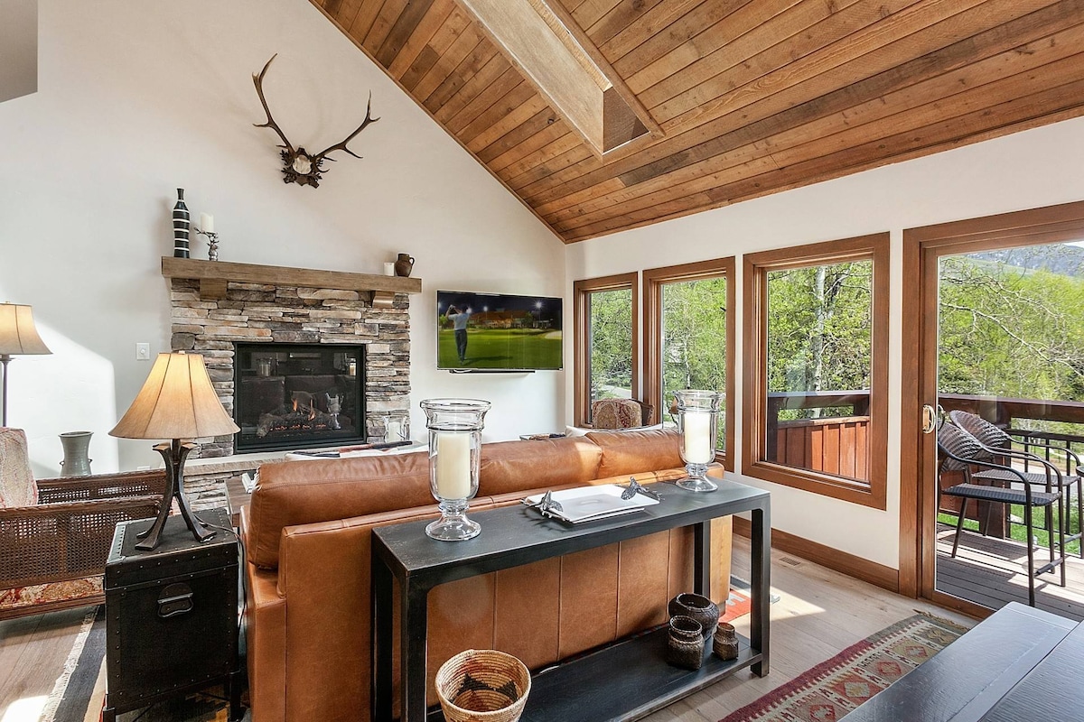 A cozy living area features a stone fireplace with a wood mantel and a flat-screen TV mounted above. Natural light floods in through large windows, revealing scenic views. Comfortable seating is arranged around a sleek coffee table, enhancing the inviting space.