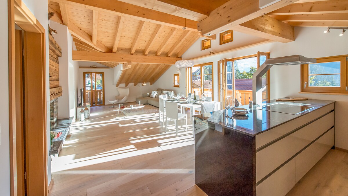 The open-plan living-dining-kitchen area features wooden beams and large windows illuminating the space. A central island kitchen is visible, with contemporary furnishings surrounding a dining table. A warm fireplace and access to outdoor balconies contribute to the inviting layout.