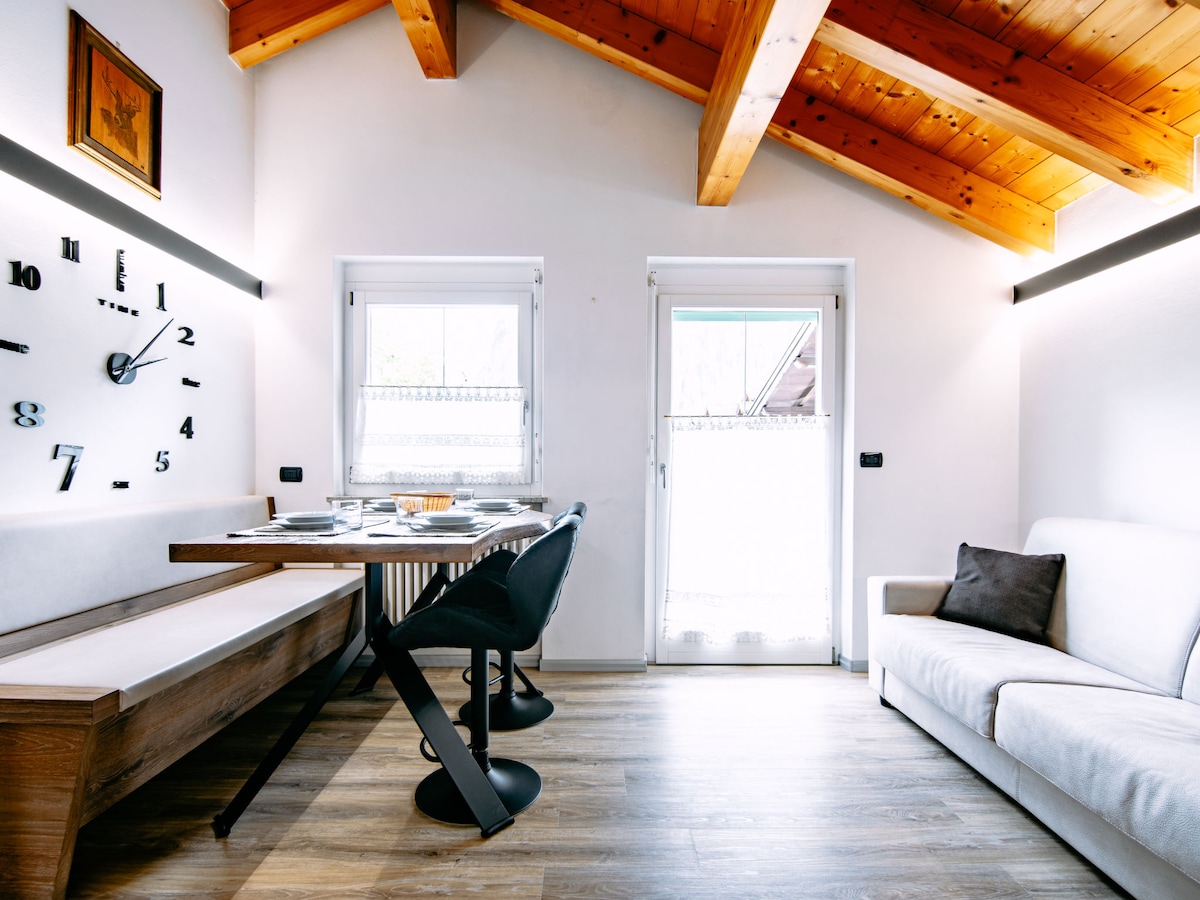 A well-lit interior features a dining area with a wooden table and four black chairs. A white sofa is positioned nearby. Large windows allow natural light, and a modern clock decorates the wall. Exposed wooden beams add a rustic touch to the space.