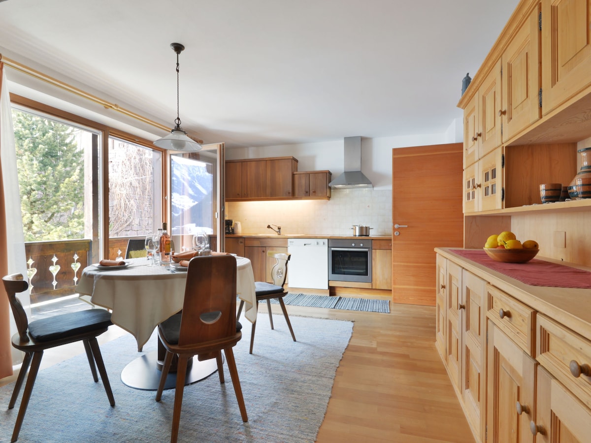An open kitchen features wooden cabinets and a dining table with four chairs. Natural light enters through large windows with a view of the outside. Modern appliances, including an oven and stove, are visible. A rug adds warmth to the parquet flooring.