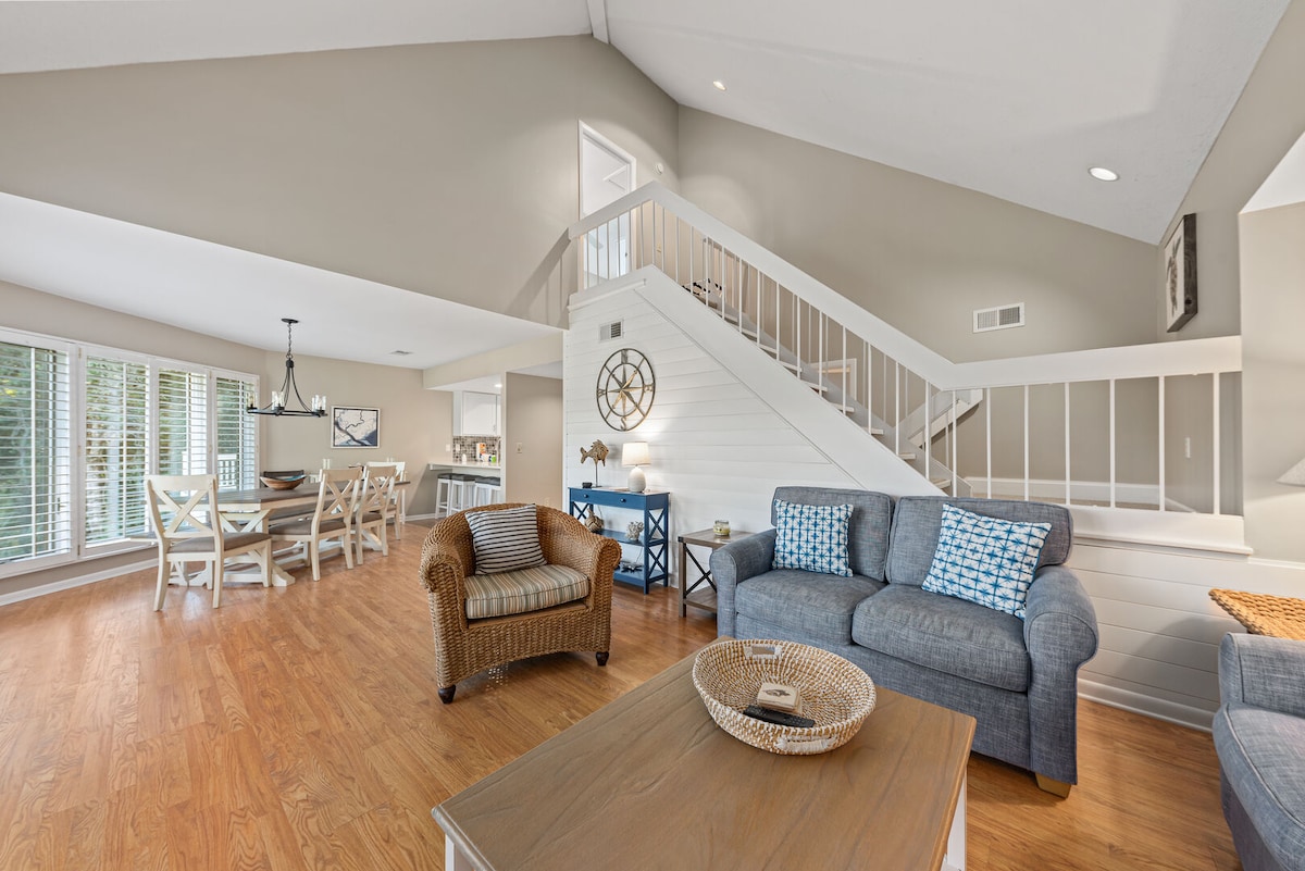 An open living area showcases a comfortable layout, featuring a gray sofa and a wicker chair. A wooden coffee table sits centrally, while a dining area with light-colored chairs is visible in the background. Large windows allow natural light to fill the space.