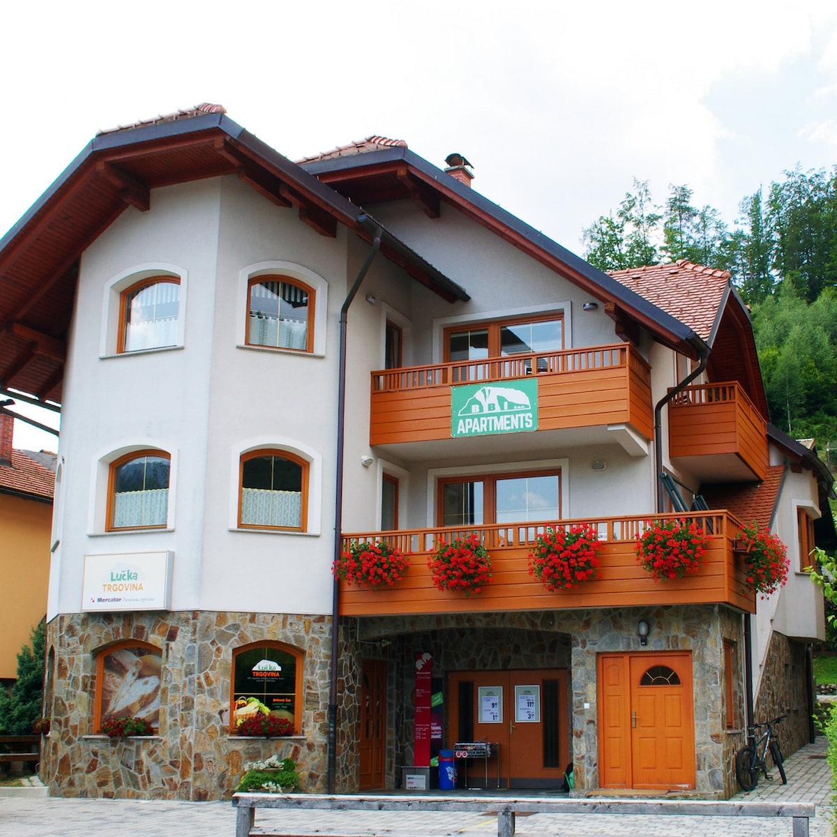 A charming building with a stone facade and wooden accents hosts the Apartments BBI. Bright flower boxes frame the windows, and a sign marking the apartments is prominently displayed. A welcoming entrance leads to the apartments located in the heart of the village.