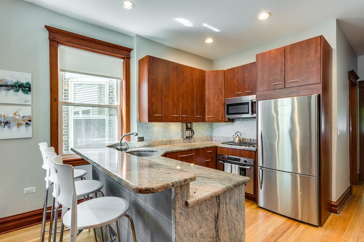 A modern kitchen features wood cabinetry and granite countertops, complemented by a stainless steel refrigerator and microwave. A spacious island is surrounded by sleek white bar stools, while natural light pours in through a large window with a roller shade.