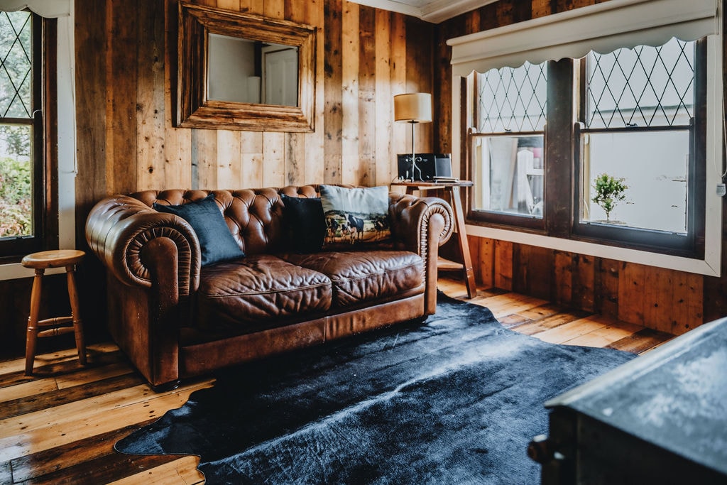 A cozy lounge area features a leather chesterfield sofa, accompanied by a wooden stool and a small side table. Natural light enters through two diamond-patterned windows, illuminating the warm wood paneling and polished wooden floor. A dark area rug adds contrast to the space.