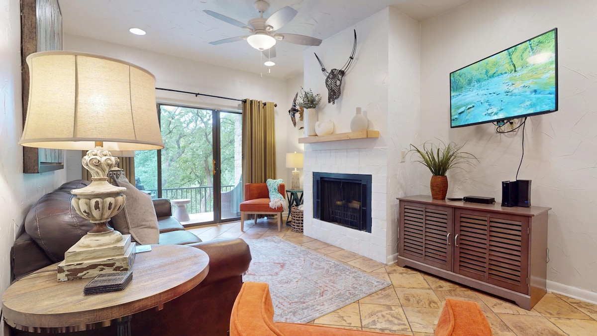 The living area features a comfortable seating arrangement with a leather sofa and accent chairs. A wall-mounted TV is positioned above a decorative fireplace. Natural light filters through large glass doors, leading to a balcony with views of the surrounding trees.