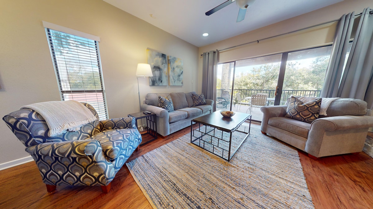 A spacious living area is furnished with two comfortable sofas and an armchair, arranged around a central coffee table. Large windows provide natural light and lead to a balcony, revealing a view of greenery outside. Neutral tones enhance the inviting atmosphere.