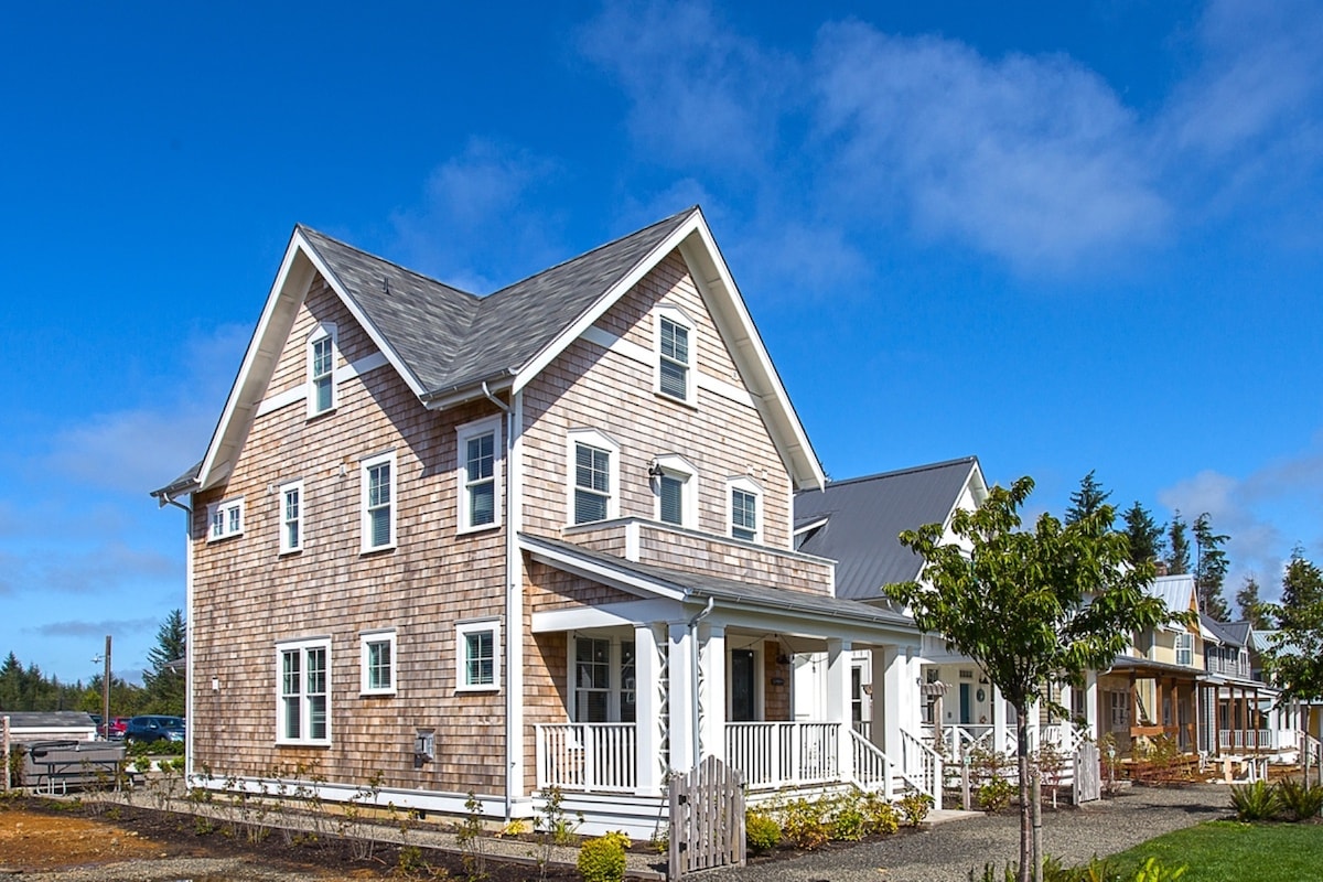 A charming three-story shingled house is set against a clear blue sky. Its multiple windows and porch provide an inviting entrance. Surrounding landscaping features fresh greenery and a gravel pathway, leading to neighboring structures that enhance the community feel.
