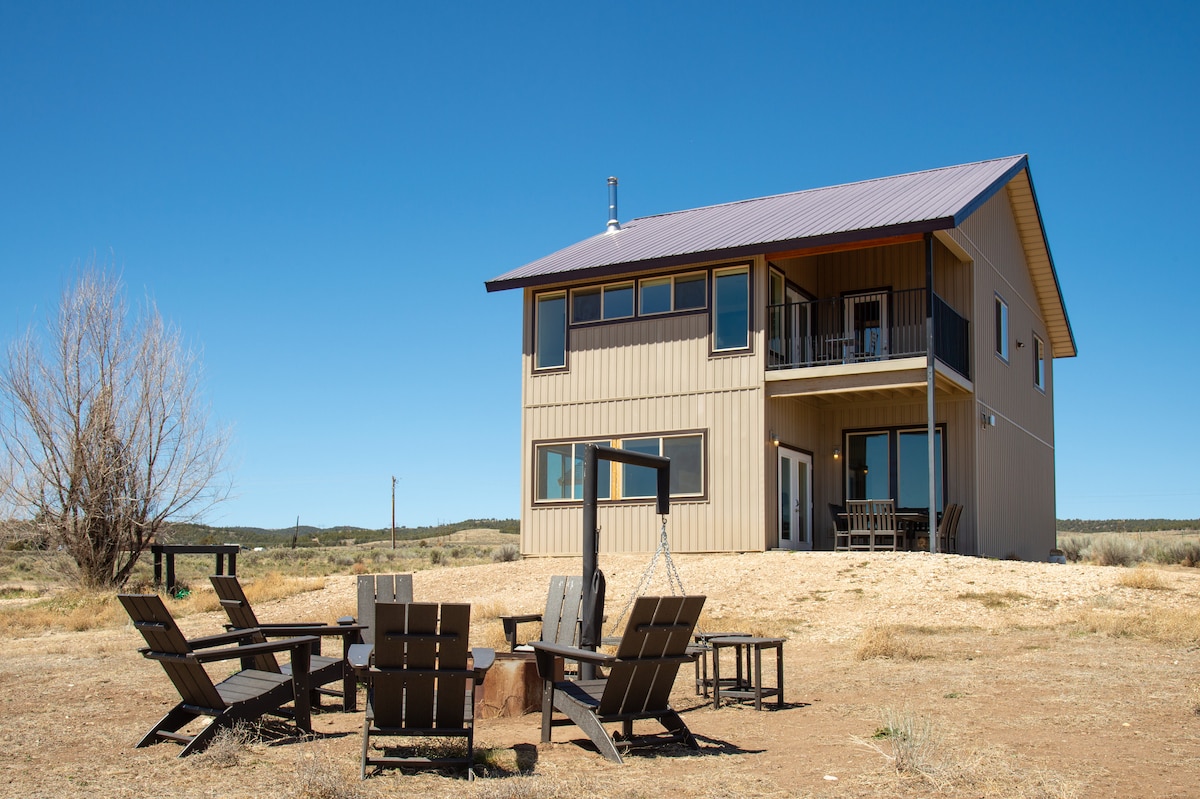 A two-story house with a modern design is illustrated against a clear blue sky. Outside, a circular arrangement of wooden seating surrounds a fire pit, providing a cozy gathering spot. The property is set amidst open land, enhancing the sense of space.