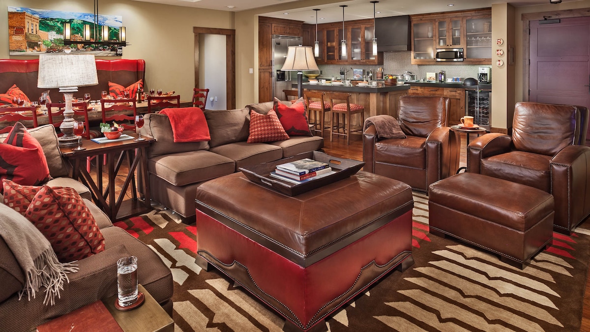 A spacious living area is arranged with comfortable brown leather sofas and chairs, complemented by decorative cushions. A stylish coffee table sits atop a patterned rug, while a well-equipped kitchen is visible in the background, featuring wooden cabinetry and modern appliances.