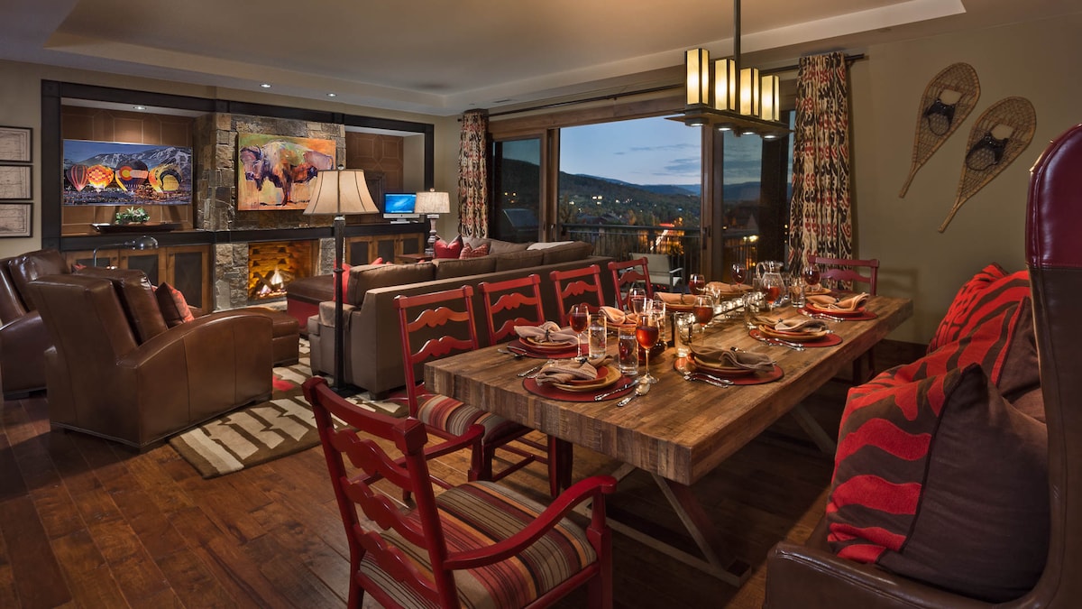 A large dining table is set for ten, surrounded by rustic seating and accented with warm decor. The open living area features a stone fireplace, comfortable seating, and expansive windows showcasing valley views. Rich wooden flooring complements the cozy atmosphere.