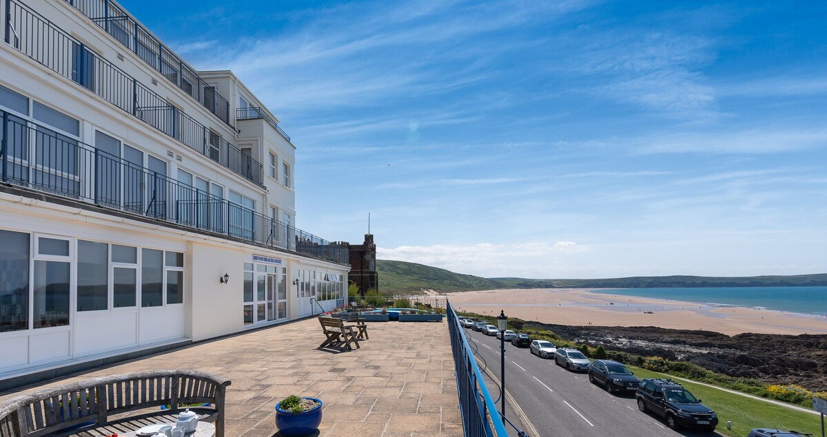 A shared terrace features seating, offering expansive views of the coastline and beach. The surrounding area displays a pleasant paved surface and a landscaped garden, with a glimpse of a road lined with parked cars below. The distant hills provide a scenic backdrop under a clear blue sky.