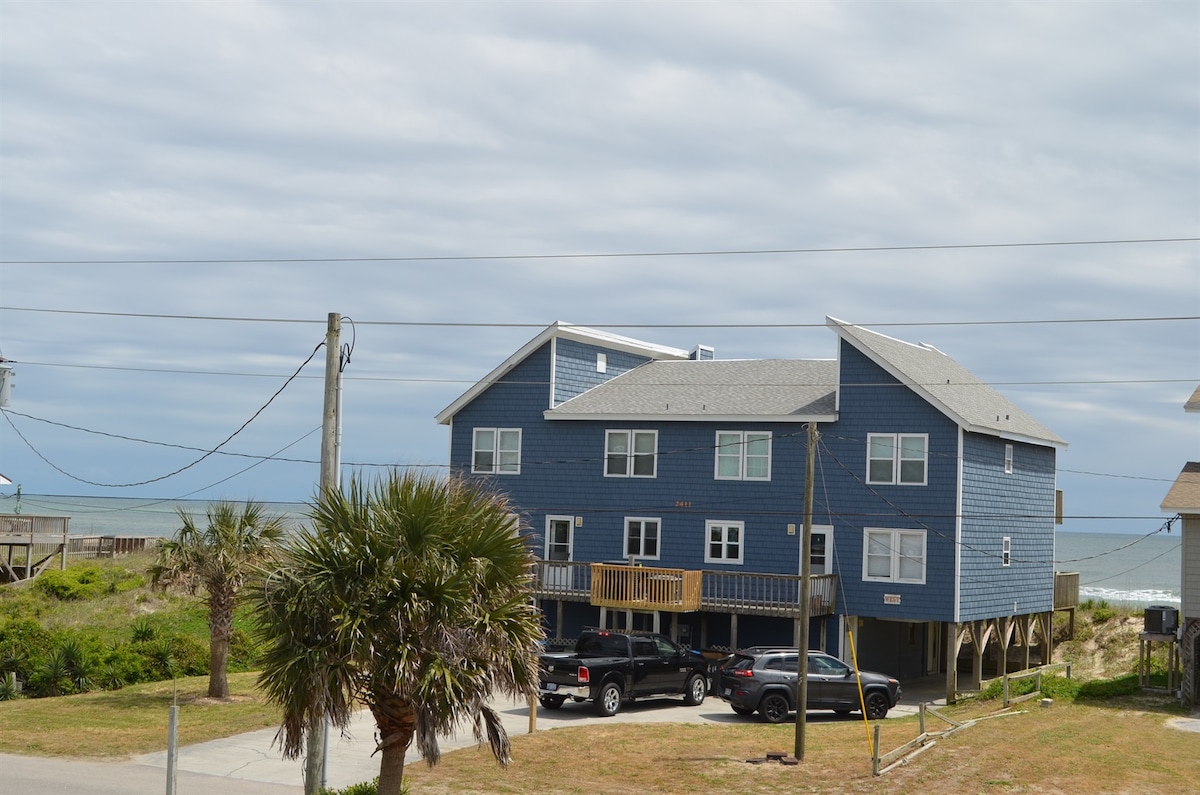 A two-story blue beach house is visible, elevated on stilts with multiple large windows. A grassy area in front contains a palm tree and space for parking several vehicles. The ocean can be seen in the distance, under a clouded sky.