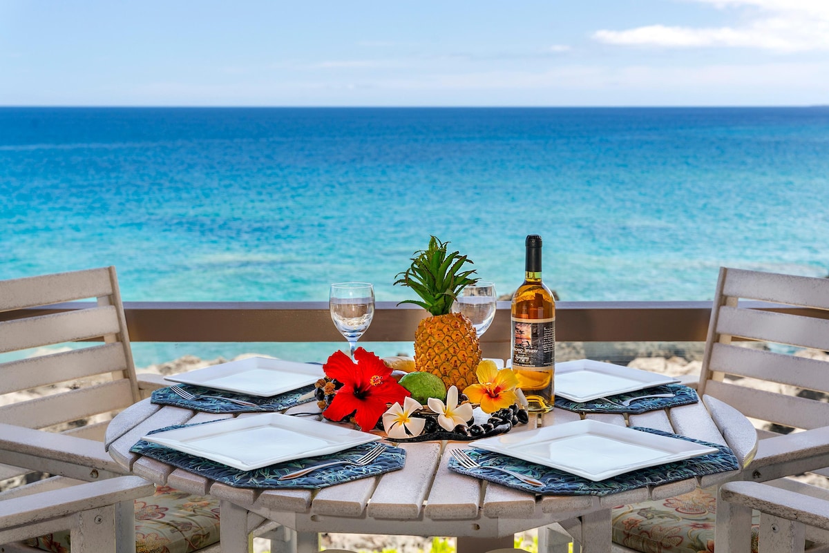 A dining table is set for four, featuring white plates and elegant glassware. A centerpiece of tropical fruits, including a pineapple and sliced lime, is surrounded by vibrant red hibiscus flowers. The ocean view in the background showcases a range of blue hues under a clear sky.