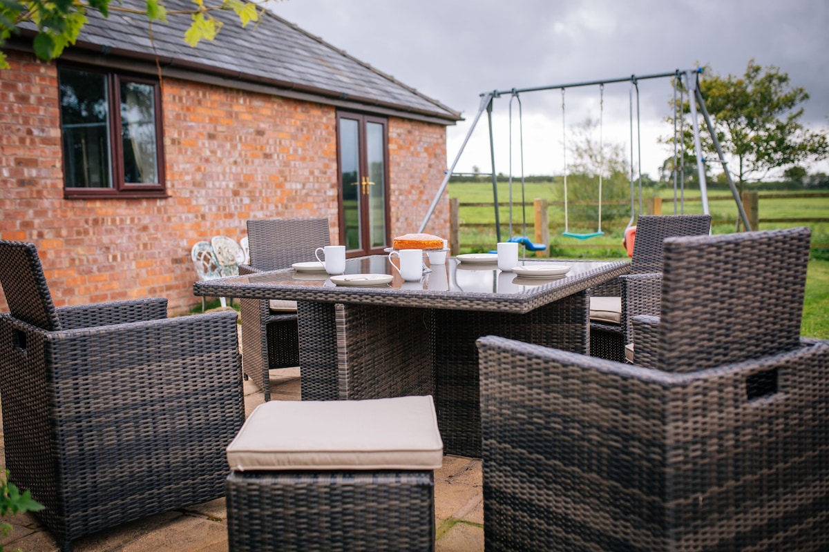 An outdoor dining area features a large table surrounded by comfortable wicker chairs, perfect for meals or relaxing. A swing set is visible in the background, with expansive fields contributing to the serene rural setting.