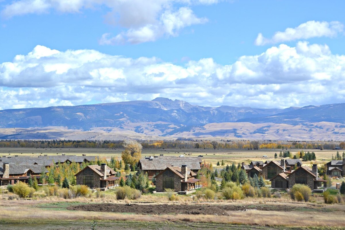 A view of the surrounding landscape shows a collection of rustic buildings nestled among trees, with expansive fields leading to distant mountains. Fluffy white clouds scatter across the blue sky, creating a serene backdrop in the Teton Village area.