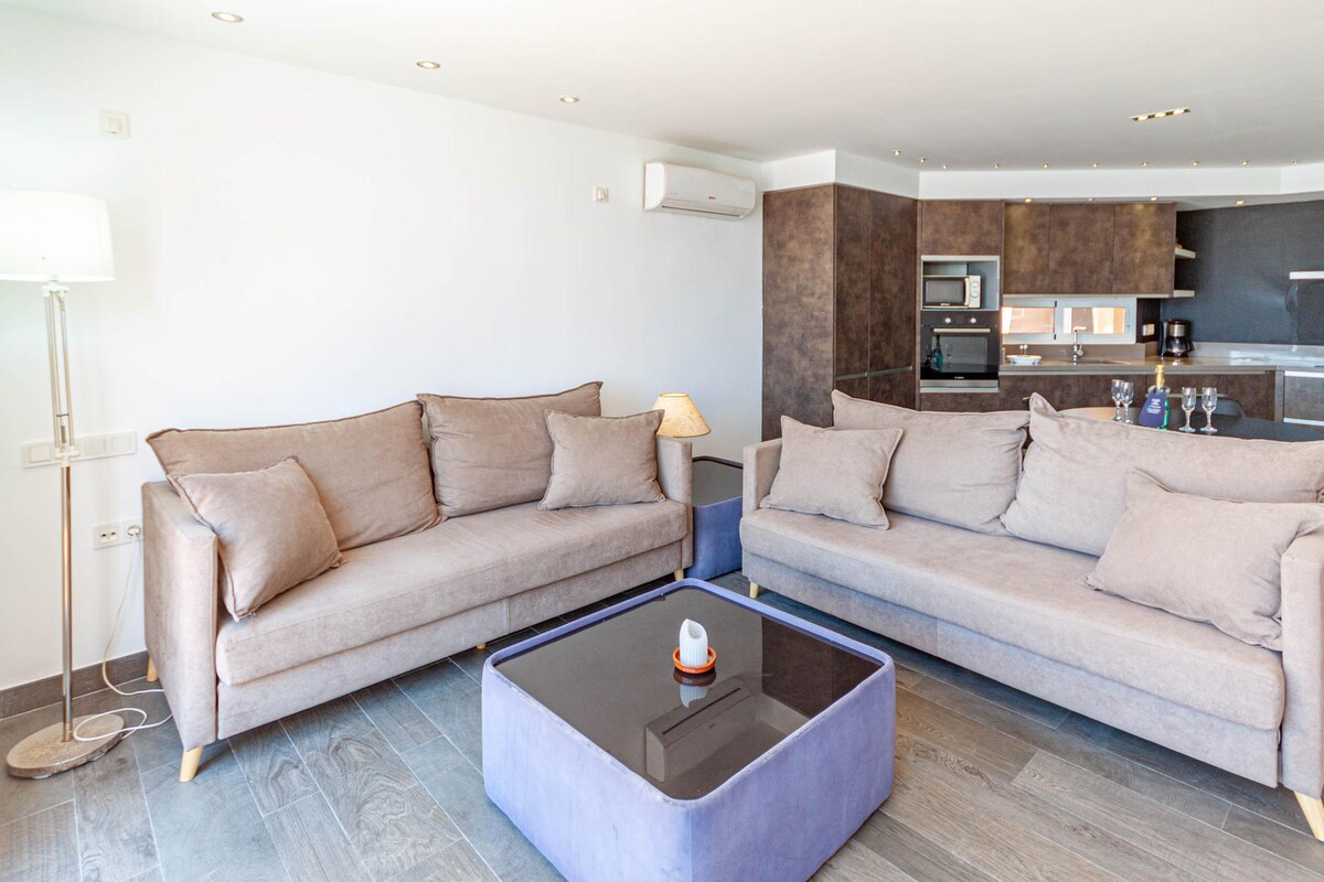 A bright and airy living room features two spacious grey sofas, arranged around a sleek dark coffee table. An accent lamp stands beside a wall, and a modern kitchen area is visible in the background, equipped with appliances and a counter.
