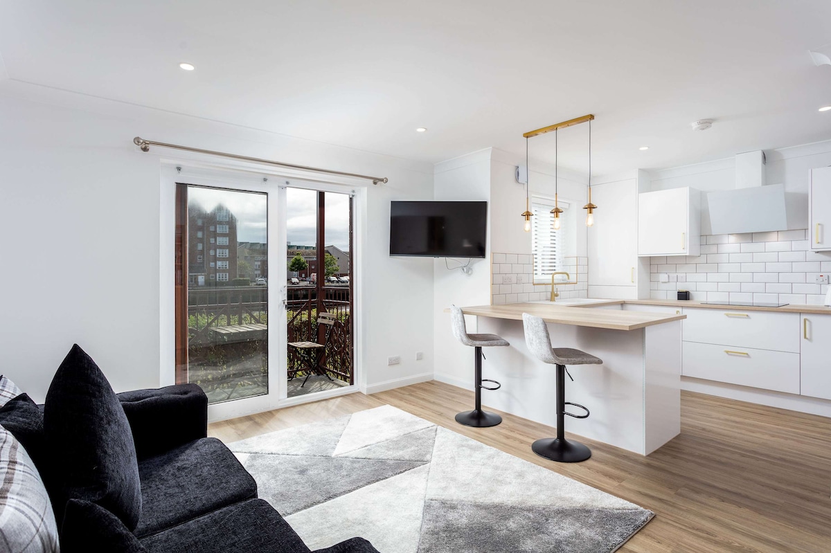 A bright and airy living space showcases an open-plan layout, with a comfortable sofa facing a wall-mounted television. A modern kitchen features white cabinetry and pendant lighting above a dining area with two bar stools, while large doors allow natural light to fill the room.