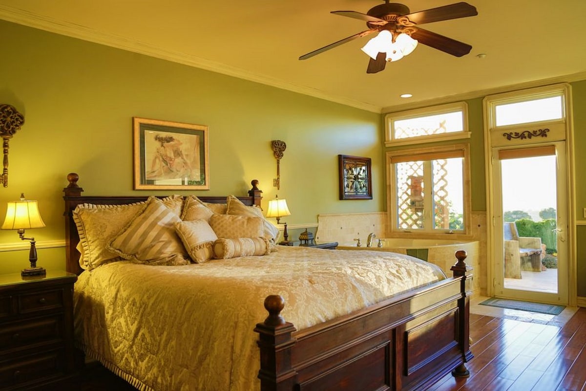 A spacious bedroom features a large bed with an elegant golden coverlet and several decorative pillows. Two bedside lamps illuminate the room, while large windows provide natural light and a view of the outdoor area. A door leads to a private patio.