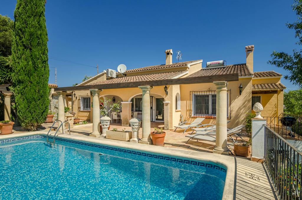 The exterior of the villa is highlighted by a private pool with a tiled border and steps leading in. Surrounding the pool, a terrace is furnished with sun loungers and potted plants. The villa features a covered outdoor sitting area, with large windows reflecting the sunny weather.